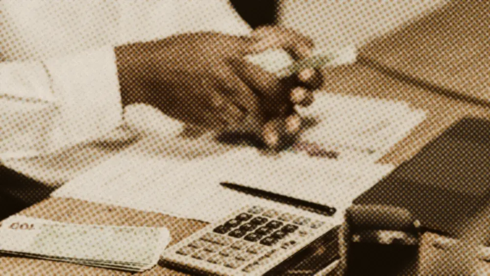 A person counting euro banknotes at a wooden desk beside a calculator, pen and financial paperwork
