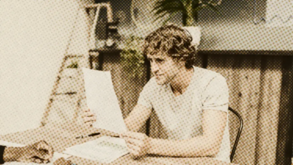 Young office worker at a wooden desk reading a document, with paperwork and a colleague across the table