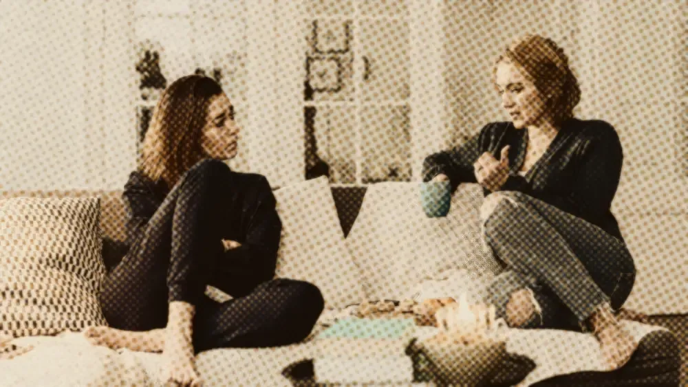 Two women in a calm, attentive conversation on a sofa in a softly lit living room, one listening with a mug of tea