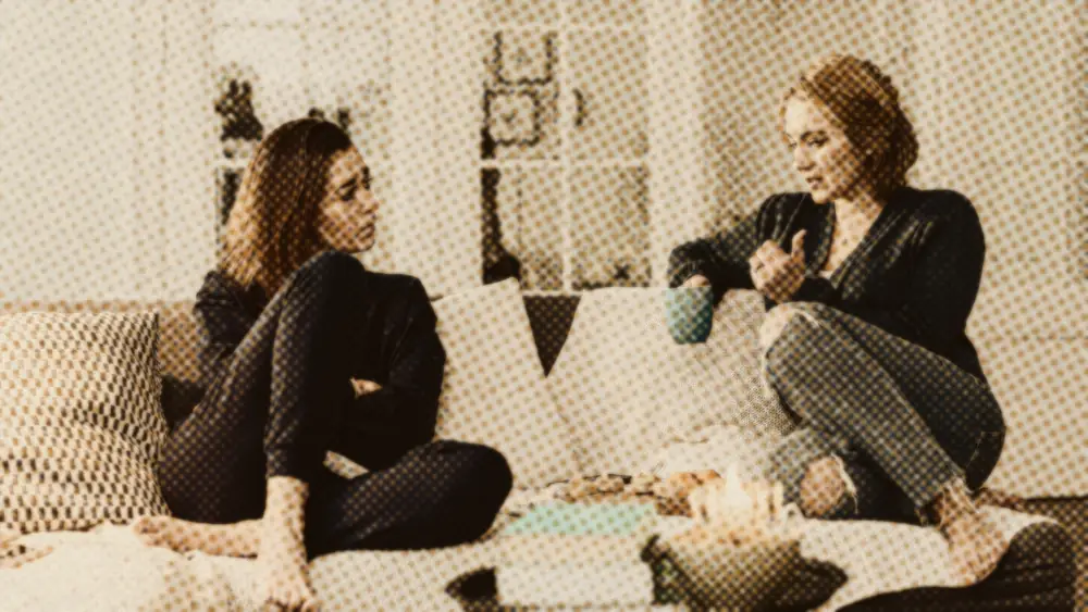 Two women in a calm, attentive conversation on a sofa in a softly lit living room, one listening with a mug of tea