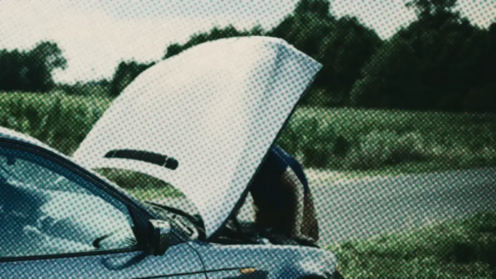 A car stopped at a country roadside with its bonnet raised, a person mostly hidden behind it.