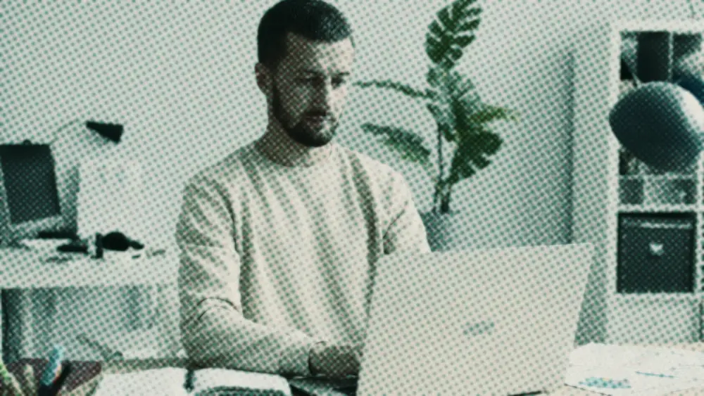Calm man at a tidy home desk reviewing on a laptop, an open notebook in front of him