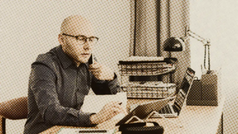 Man in glasses at a wooden desk holding a phone to his ear, a white sheet of paper in his other hand