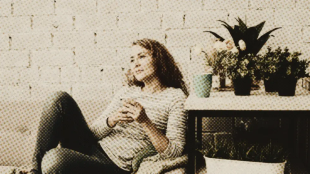 Calm woman on a sofa holding her phone and gazing up thoughtfully, taking a considered pause in a warm sunlit room