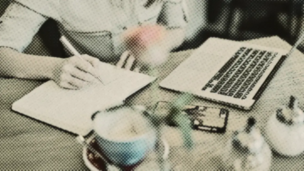 Hands writing monthly figures in an open notebook beside a laptop and coffee on a warm wooden table