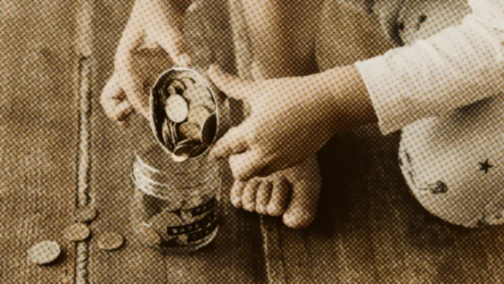 A child carefully tips a small tin of coins into a glass savings jar on a warm wooden floor