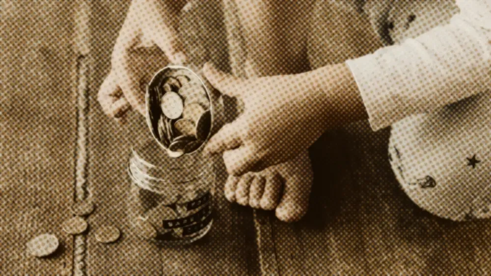 A child carefully tips a small tin of coins into a glass savings jar on a warm wooden floor