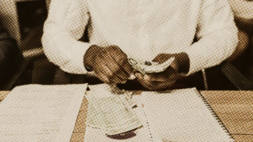Person at a desk counting 100-euro banknotes beside a handwritten budget notebook and household papers
