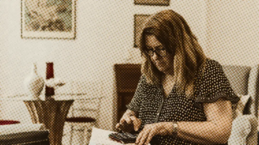 An older woman in glasses sits at home using a calculator and notebook to work through her household budget.