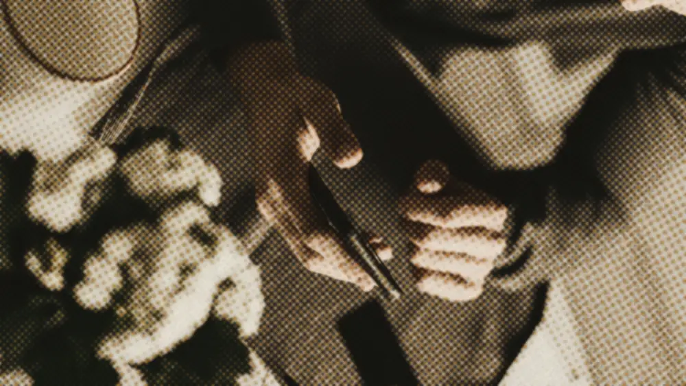 A man's hands at a quiet desk, one cradling a dark-screened phone, the other resting, beside a cup and flowers