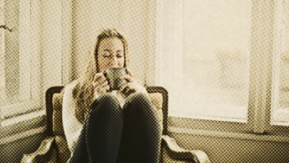 Woman in a cosy armchair by a bright window, eyes closed, holding a warm mug to her face