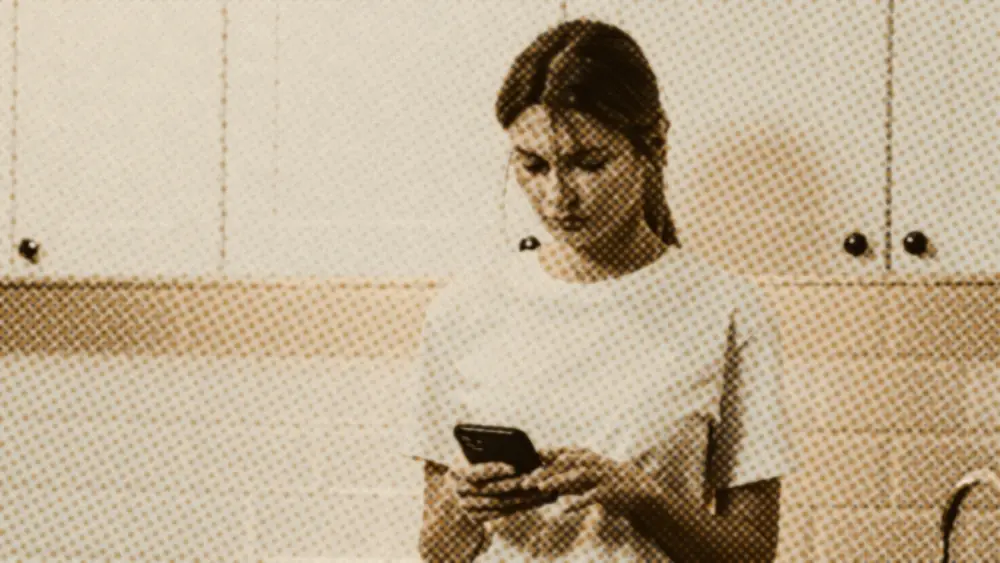 A young woman standing in a kitchen looking down at her phone with a quiet, pensive expression