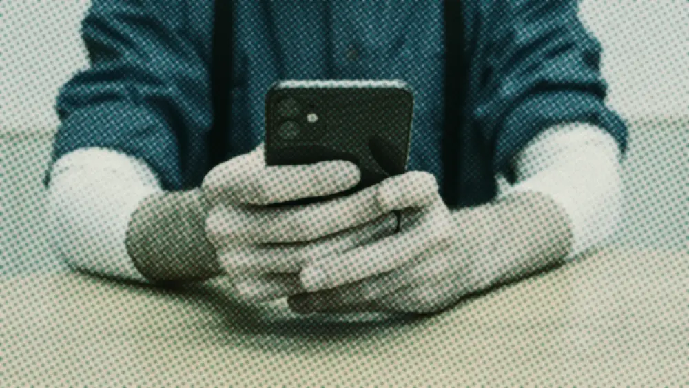 A person in a blue shirt holds a smartphone in both hands above a plain wooden table