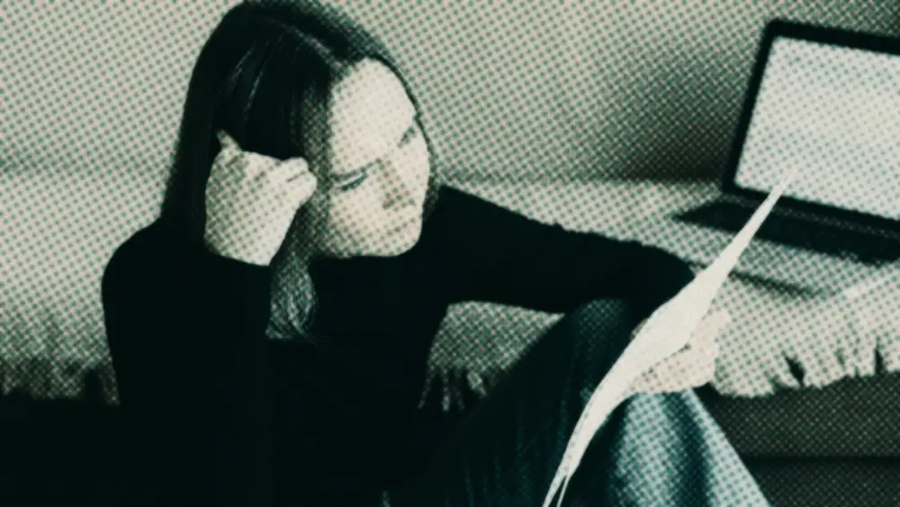 Woman in a black jumper reading a paper statement, hand to her temple, an open laptop beside her at home