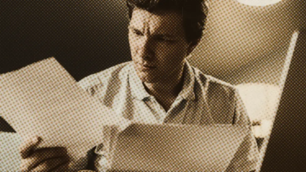 A man at a warmly lit desk frowns as he reads a printed statement beside an open laptop