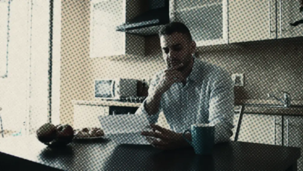A man at a sunlit kitchen table, hand on chin, reading a letter beside a coffee mug, weighing it over