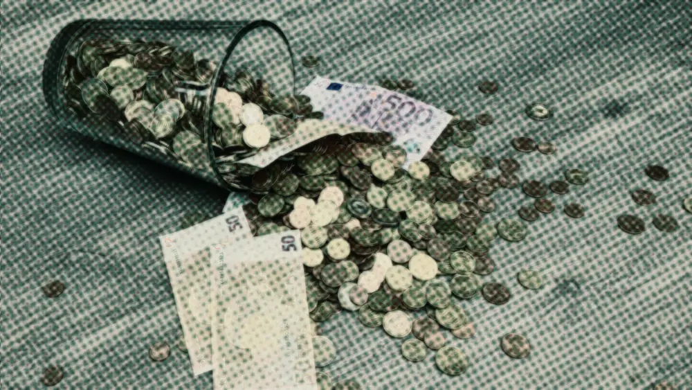 A glass jar tipped on its side spilling euro coins and banknotes across a wooden floor