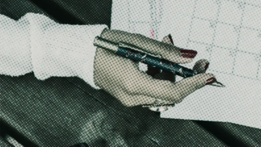 A hand holds a pen over a printed monthly calendar on a wooden table, poised to mark dates