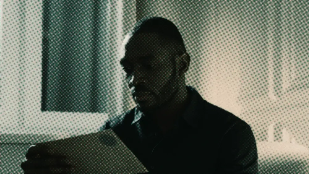 A man sits alone by lamplight at night, quietly reading a letter and a stack of opened envelopes