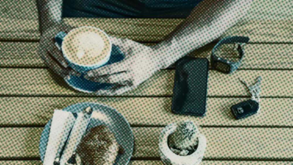 Hands cradling a cappuccino beside a blank phone screen, car keys and a croissant on a slatted wooden table