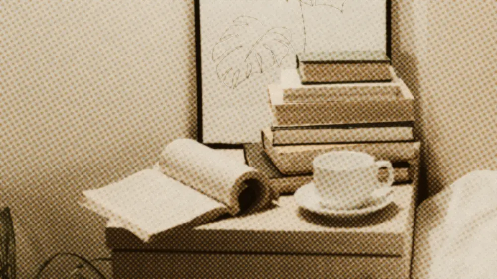 White cup and saucer beside a face-down open book on a bedside cabinet, stacked books and a bed behind