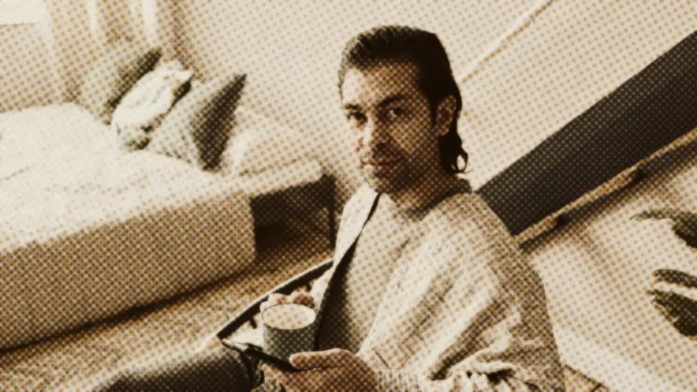 Calm man holding a mug of coffee at home, sitting composedly beside his bed and looking to camera
