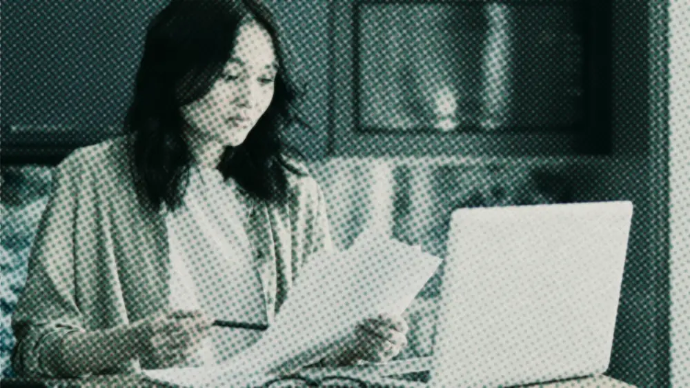 Young woman at a home table calmly reading a printed fund document, a laptop and notebook beside her