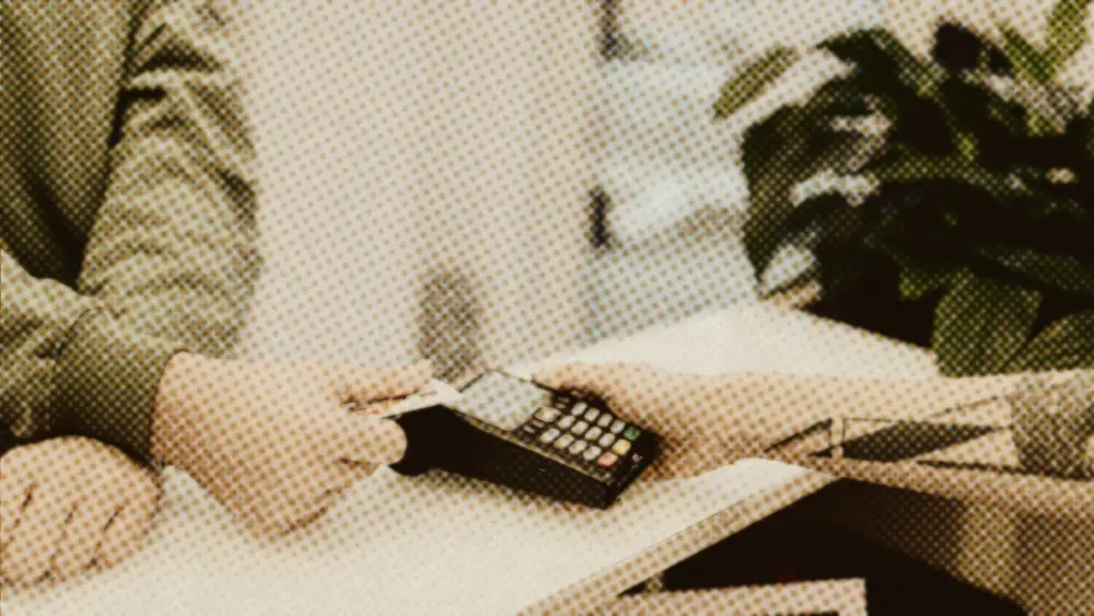 Customer taps a bank card on a shop payment terminal held by a worker across a counter, a leafy plant behind