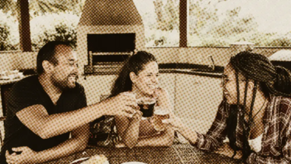 Three friends in their thirties laughing and clinking glasses over a shared meal at an outdoor table