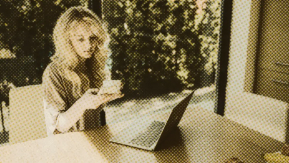 Calm woman reviewing her phone beside an open laptop at a sunlit kitchen table