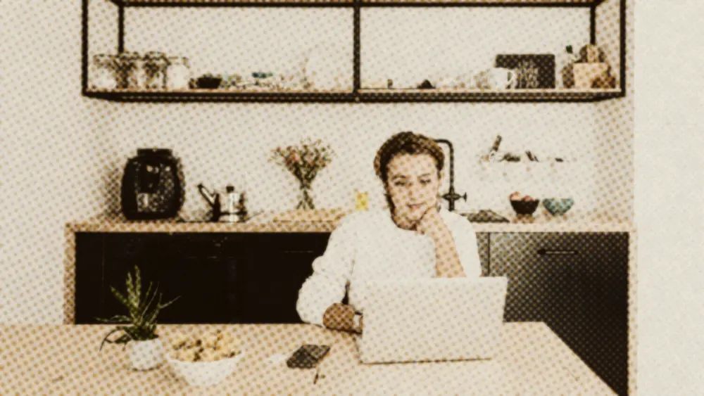 Calm woman at a sunlit kitchen table, chin resting on her hand, focused on her laptop in a tidy modern kitchen
