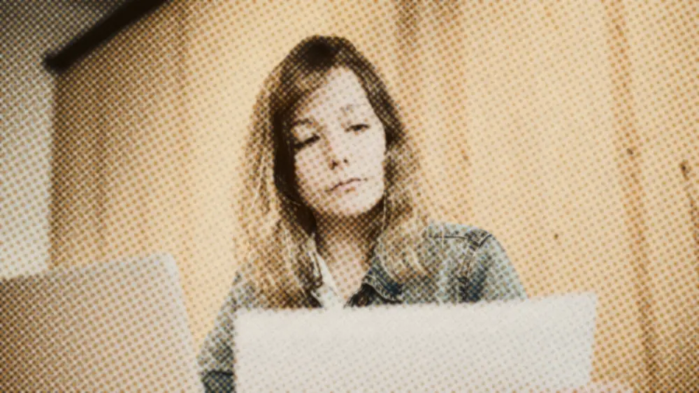 Woman in a denim jacket holding up a printed document to read, a laptop beside her against a wood wall