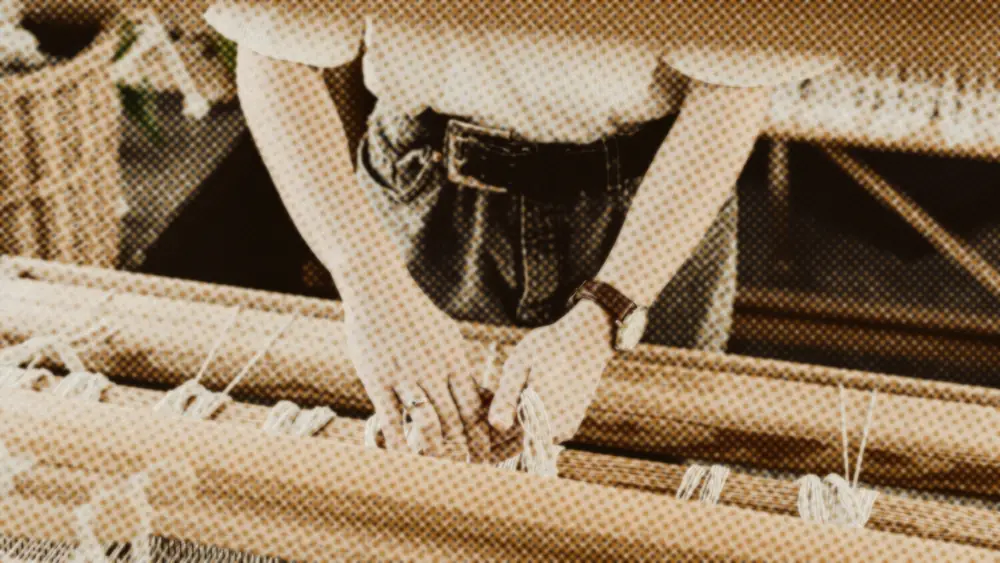 Craftswoman leaning over a wooden loom, knotting warp threads by hand in her workshop