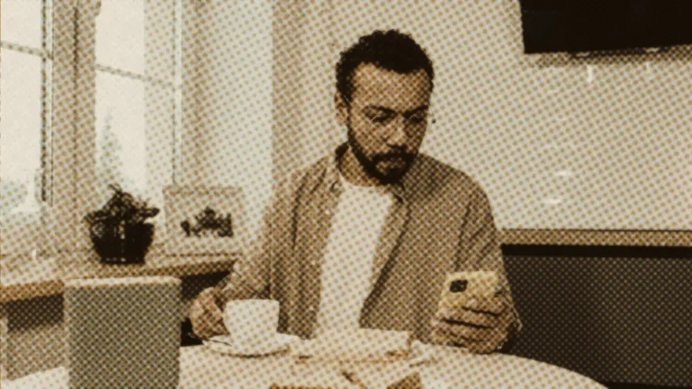 Calm man at a sunlit kitchen table looking at his phone beside a cup of coffee